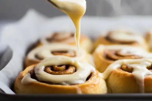 A tray of cinnamon rolls being frosted with cream cheese icing while steam rises from the freshly baked rolls