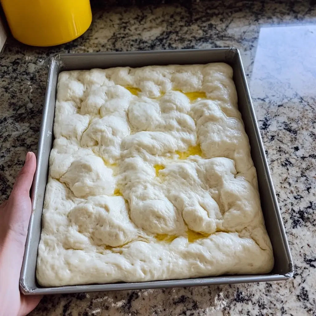 A tray of sourdough discard focaccia dough with visible dimples created by hand, glistening with olive oil, placed on a granite countertop.