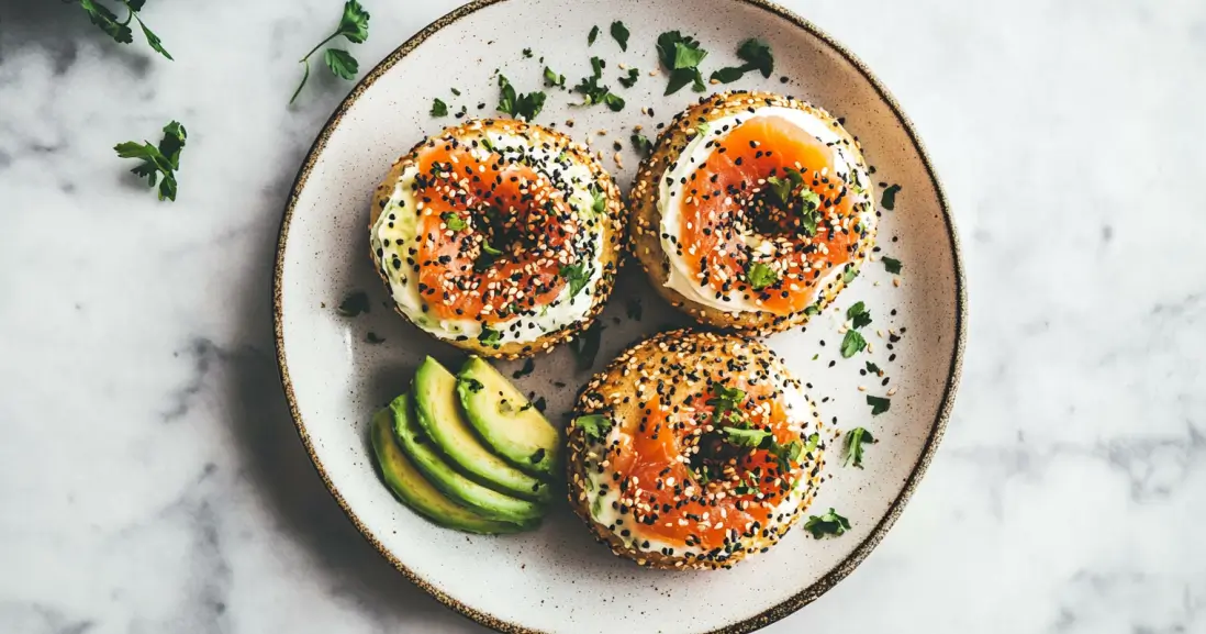 Three sourdough discard bagels topped with cream cheese, smoked salmon, and seeds, served with sliced avocado on a ceramic plate